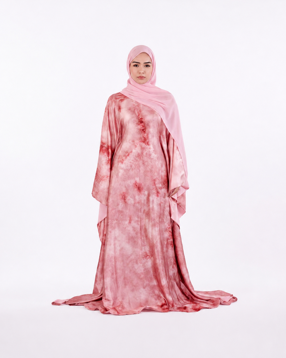 A woman wearing a long pink abaya with a red tie-dye pattern and a matching light pink hijab, standing against a seamless white studio background.