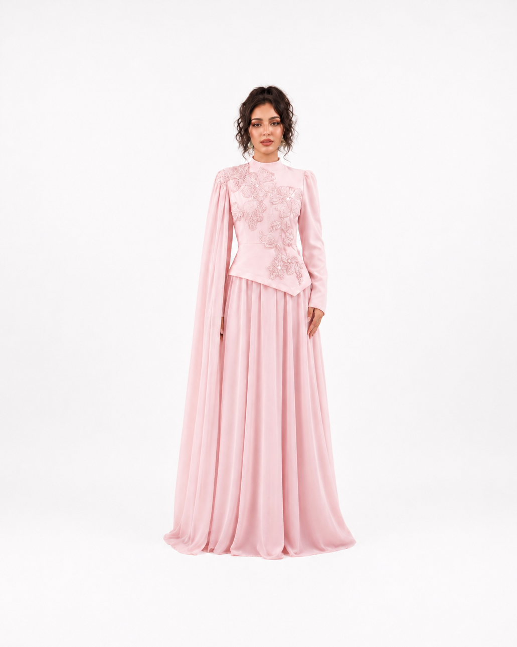 A woman modeling a long pink formal gown with a high neck, sheer draped cape sleeves, and intricate floral embroidery on the bodice, set against a plain white studio background.
