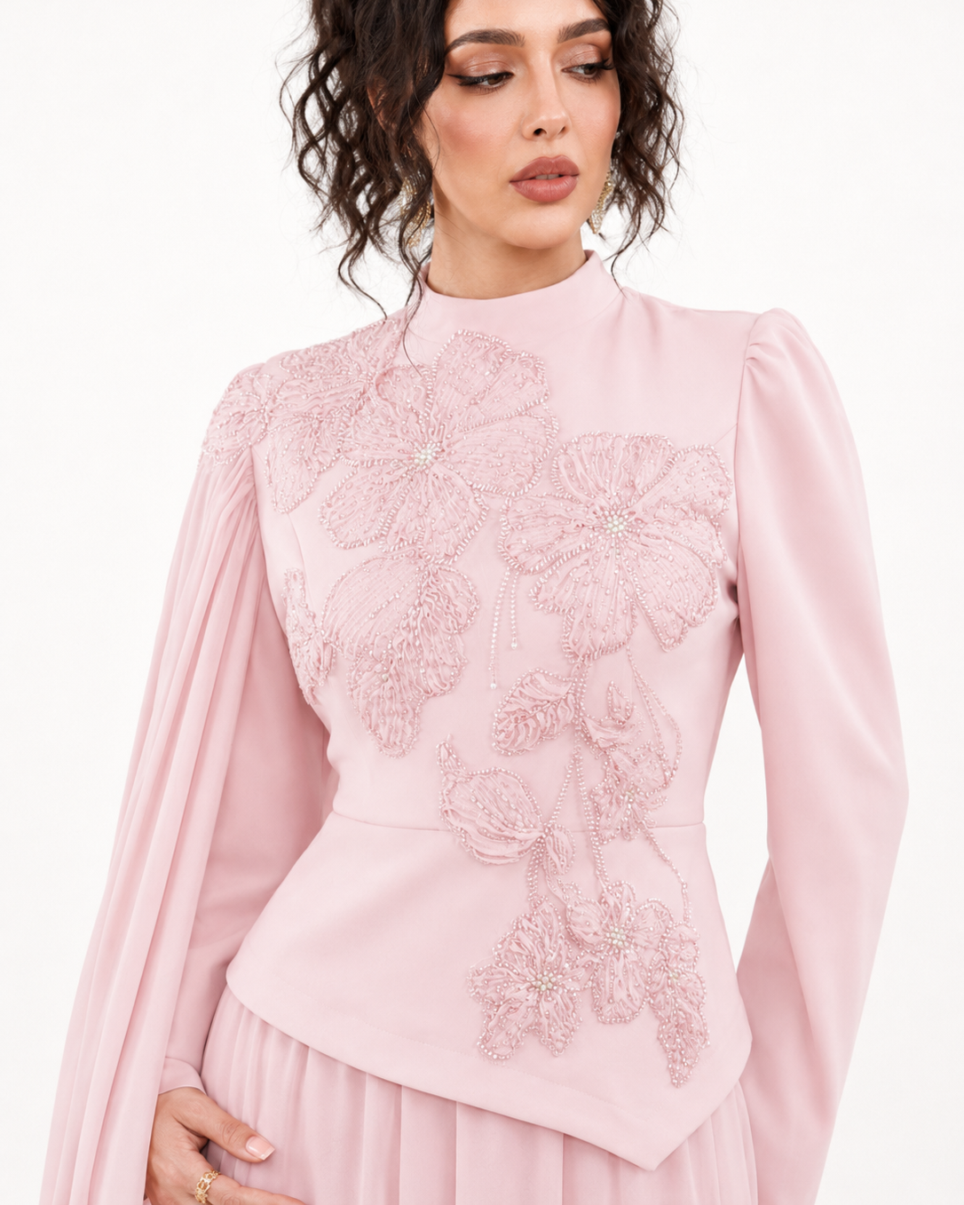A woman wearing a light pink high-neck blouse with long sheer sleeves and large floral applique embroidery on the front, paired with a matching pleated skirt against a plain white background.