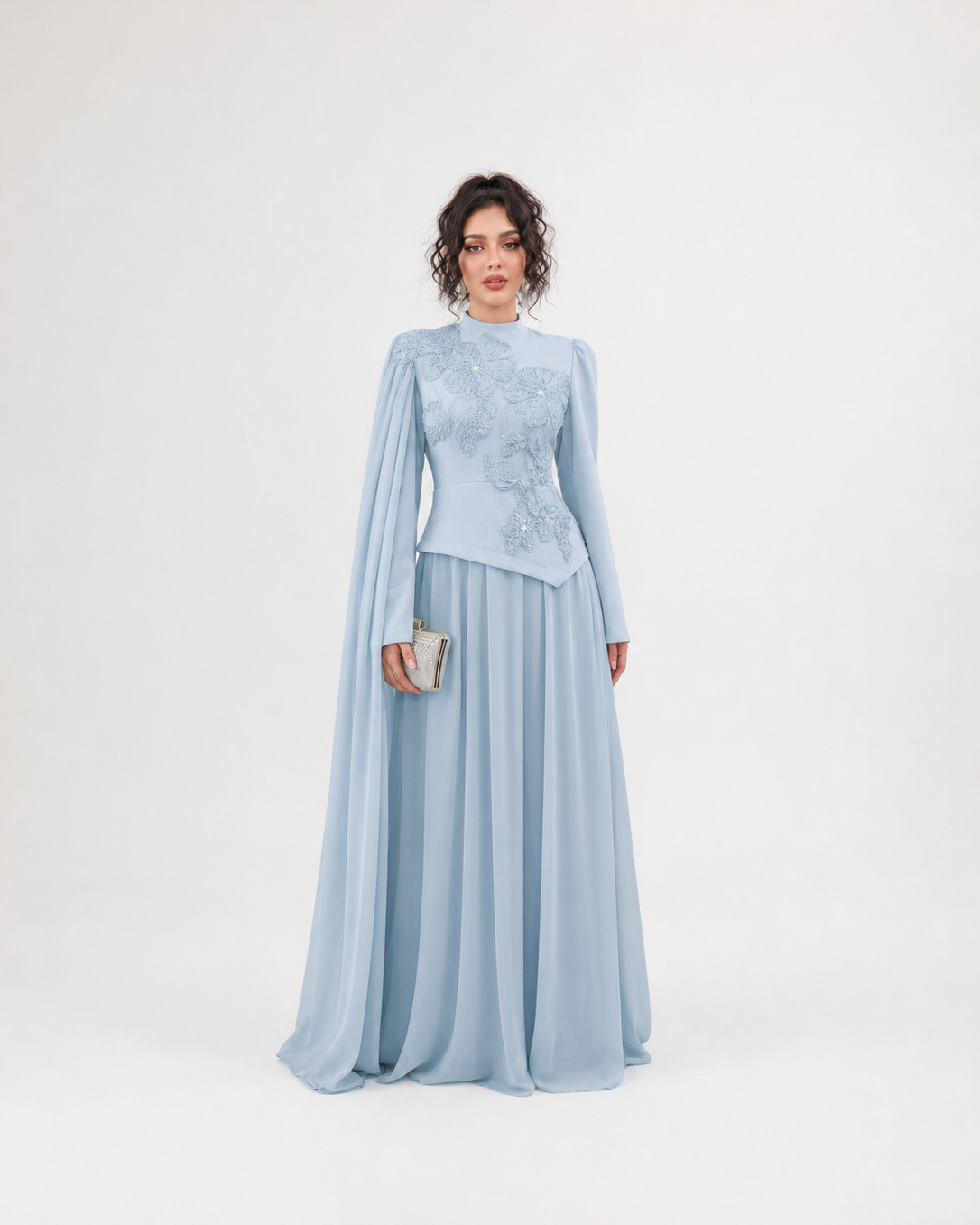 A woman modeling a light blue formal dress with a high mock neck, long draped cape sleeves, and intricate floral embroidery on the bodice, set against a plain white studio background.