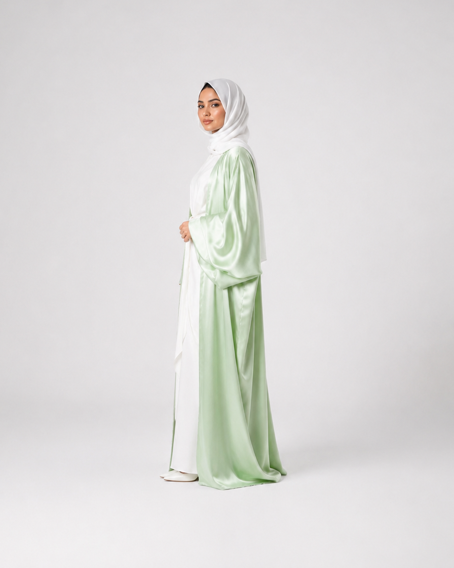 A woman wearing a light sage green and white satin abaya with a matching white hijab, standing in profile against a light gray studio background.