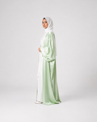 A woman wearing a light sage green and white satin abaya with a matching white hijab, standing in profile against a light gray studio background.