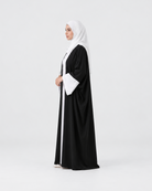 A woman wearing a black abaya with white trim and a white hijab, standing in profile against a light gray studio background.