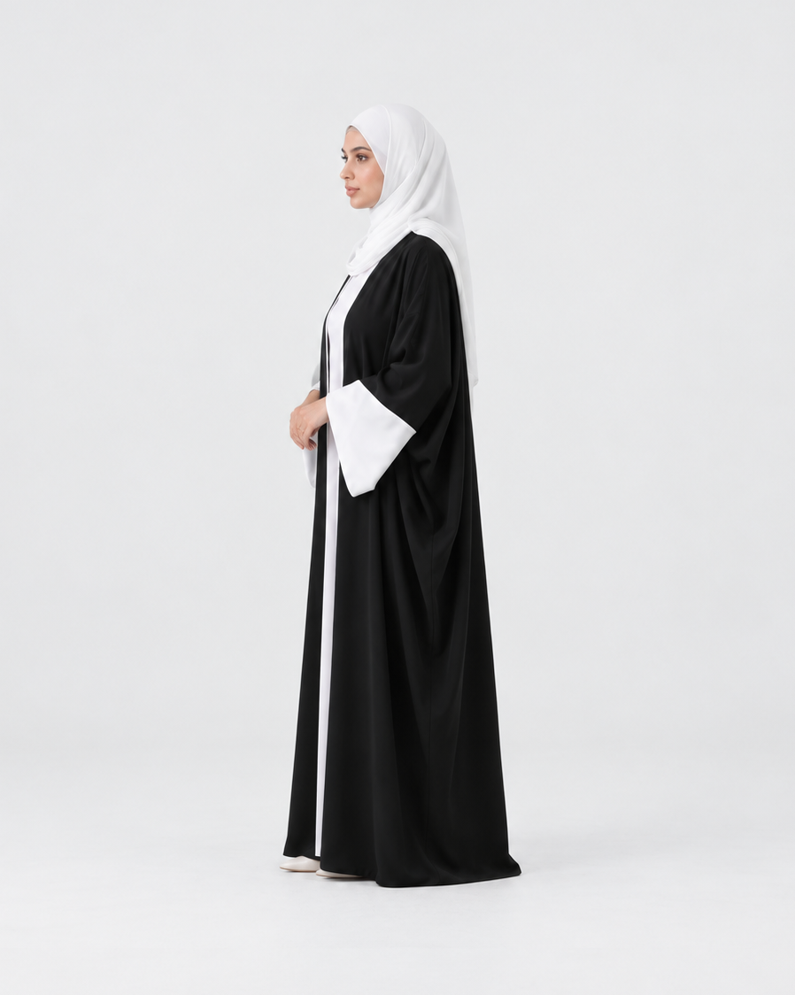 A woman wearing a black abaya with white trim and a white hijab, standing in profile against a light gray studio background.