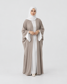 A woman modeling a modest outfit consisting of a taupe-gray abaya with white inner dress and a white hijab, standing against a seamless light gray studio background.