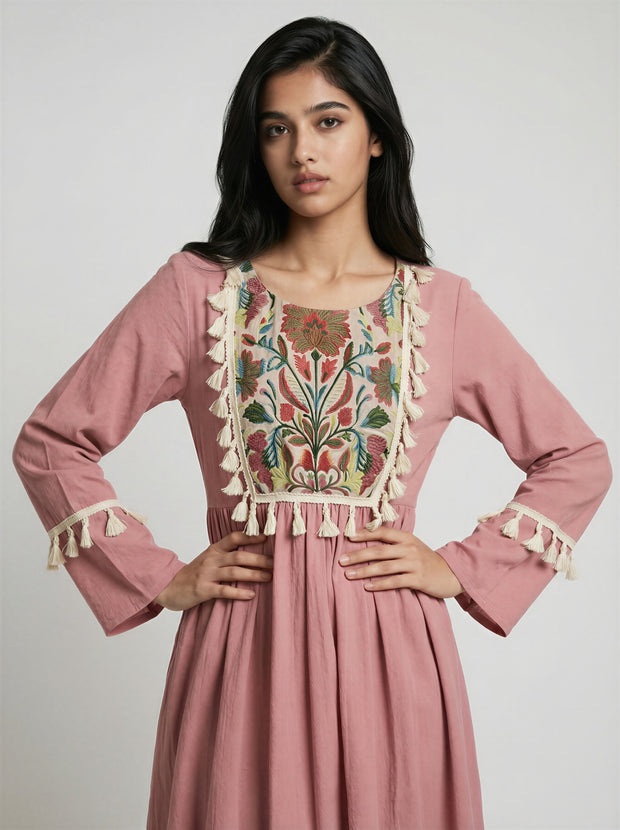 Woman wearing a pink dress with intricate embroidery and tassel details on a plain background