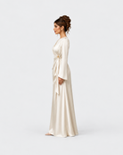 A woman in profile wearing a floor-length cream satin gown with long sleeves and a self-tie waist, photographed against a seamless light gray studio background.