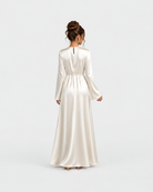 A woman viewed from behind wearing a floor-length satin gown in a warm ivory or cream tone, standing against a clean light gray studio background.
