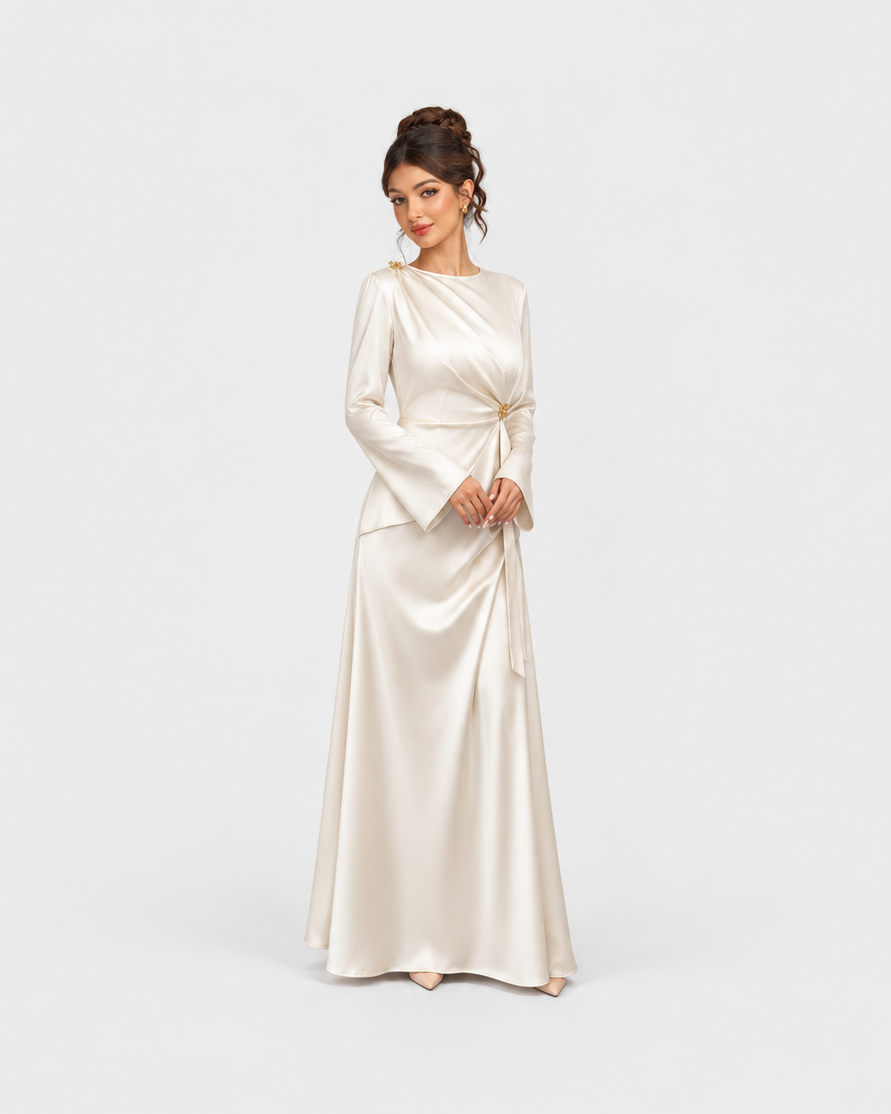 A woman modeling a floor-length satin gown in a warm ivory or cream shade against a plain light gray studio background. The dress has long sleeves, a draped neckline, and a self-tie waist detail.