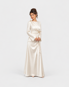 A woman modeling a floor-length satin gown in a warm ivory or cream shade against a plain light gray studio background. The dress has long sleeves, a draped neckline, and a self-tie waist detail.