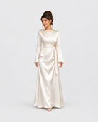 A woman modeling a floor-length satin gown in a warm ivory or cream shade against a light gray studio background. The dress has long sleeves, a draped neckline, and a self-tie waist sash.