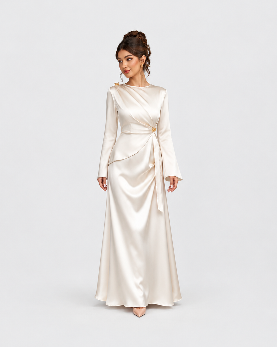 A woman modeling a floor-length satin gown in a warm ivory or cream shade against a light gray studio background. The dress has long sleeves, a draped neckline, and a self-tie waist sash.