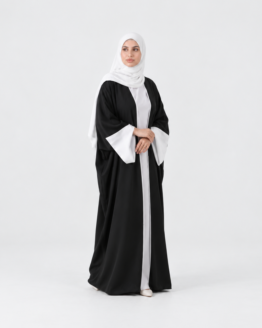 A woman modeling a black abaya with wide white bell sleeves and a white inner layer, paired with a white hijab, standing against a seamless light gray studio background.
