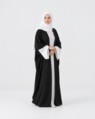 A woman modeling a black abaya with wide white bell sleeves and a white inner layer, paired with a white hijab, standing against a seamless light gray studio background.